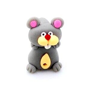 Könnte beinhalten: Eine graue und rosafarbene Mausfigur mit gelbem Bauch und roter Nase. Die Maus sitzt aufrecht mit eingezogenen Pfoten.