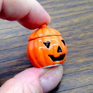 May include: A small, orange ceramic pumpkin with a lid, featuring a carved jack-o'-lantern face with black eyes and a smiling mouth. The pumpkin is held in a hand, showcasing its glossy finish and festive design, perfect for Halloween.