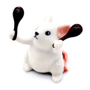 May include: A white ceramic bunny figurine holding two dark brown spoons. The bunny has pink accents on its ears and tail, with black details for the eyes and nose. The spoons are glossy and add a contrasting element to the figurine.