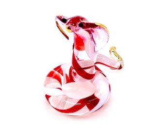 May include: A glass cobra figurine with red and white stripes. The cobra is coiled, with its head raised and hood flared. The figurine has gold accents on the hood and eyes. The snake is a decorative item.