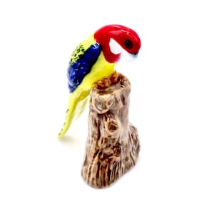 May include: A ceramic parrot figurine with a red head, yellow body, blue wings, and a white beak, perched on a brown tree stump. The stump has textured bark. A decorative item.