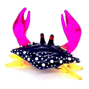 Könnte beinhalten: Glasfigur eines Krebses mit dunkelblauem Körper und weißen Punkten. Der Krebs hat leuchtend pinke Scheren und gelbe Beine. Die Augen sind schwarz mit roten Spitzen. Vor weißem Hintergrund.