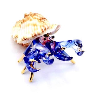Puede incluir: Figurita de cristal de un cangrejo ermitaño. El cuerpo del cangrejo es una mezcla de tonos azules y rojos, con detalles dorados en las pinzas y las patas. La concha es de color beige natural. Sobre fondo blanco.