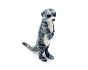 Może przedstawiać: Ceramiczna figurka surykatki w pozycji stojącej. Surykatka ma szaro-biały, nakrapiany wzór, z białym brzuchem i małym, ciemnym nosem. Figurka ma około 10 cm wysokości.