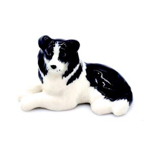 Könnte beinhalten: Eine Keramikfigur eines Hundes, hauptsächlich weiß mit schwarzen Markierungen. Der Hund befindet sich in einer entspannten, liegenden Pose, mit ausgestreckten Vorderpfoten. Die Figur hat eine glänzende Oberfläche und steht vor weißem Hintergrund.