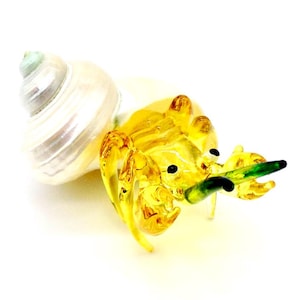 Puede incluir: Una figurita de vidrio de un cangrejo ermitaño con una concha blanca y un cuerpo amarillo. El cangrejo sostiene un alga verde en sus garras.