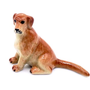 May include: A ceramic figurine of a dog in a seated position. The dog has a light brown coat with darker brown accents on the ears, nose, and paws. The figurine is detailed with black eyes and a black nose. The dog's tail is extended.