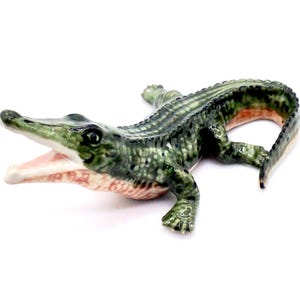 May include: A ceramic alligator figurine in shades of green, with a detailed textured body and open mouth revealing a pink interior. The figurine is posed on a white background, showcasing its realistic features and design.