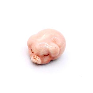 Peut inclure: Petite figurine en céramique rose représentant un porcelet endormi. Le porcelet est recroquevillé, les yeux fermés et le groin visible. La figurine a une finition brillante et est posée sur un fond blanc.
