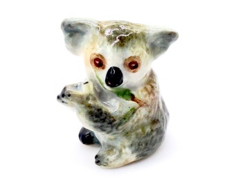Może przedstawiać: Ceramiczna figurka koali z małym koalą trzymającym się z boku. Figurka jest pomalowana w odcieniach szarości, brązu i bieli, z czerwonymi oczami i czarnym nosem. Koala trzyma zielony liść.