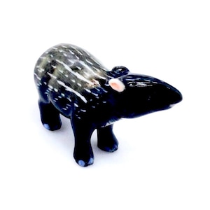 Puede incluir: Figura de cerámica de un tapir, con cuerpo negro brillante y marcas blancas y grises. El tapir tiene orejas pequeñas y redondeadas con detalles rosas y azul en las pezuñas. La figura está sobre un fondo blanco.