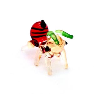 Könnte beinhalten: Eine Miniatur-Glasbienenfigur. Die Biene hat einen rot-schwarz gestreiften Körper, klare Beine und grüne und beige Flügel. Die Figur ist klein und detailliert, mit einem skurrilen Design.