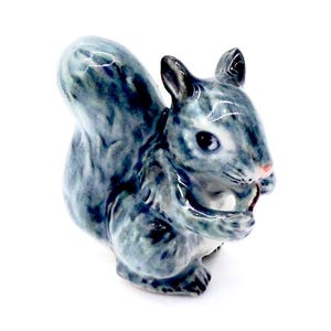 May include: A ceramic squirrel figurine in shades of blue and gray, holding a nut. The squirrel has detailed fur texture, a pink nose, and black eyes. The figurine is set against a white background.