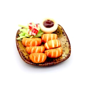 May include: A miniature ceramic plate with a brown and green glaze, featuring a small serving of orange-coloured food, a dollop of brown sauce, and a green and white salad.