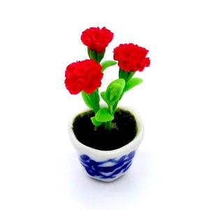 May include: Miniature potted plant with three vibrant red carnation flowers and green leaves. The small ceramic pot is white with a blue floral design. The pot is filled with dark soil, creating a charming, decorative piece.