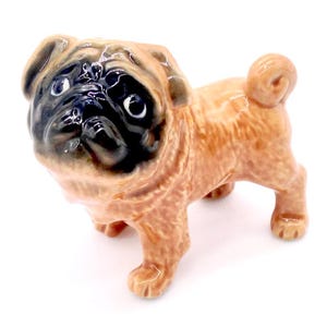May include: A small ceramic figurine of a pug dog. The pug is brown and has black facial features. The figurine is standing on all four legs and has its tail curled up.
