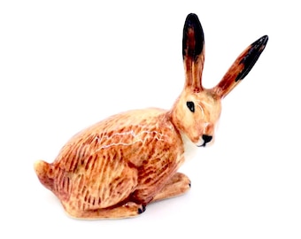 May include: A small ceramic figurine of a brown hare sitting with its ears up. The hare is detailed with brown and white fur.