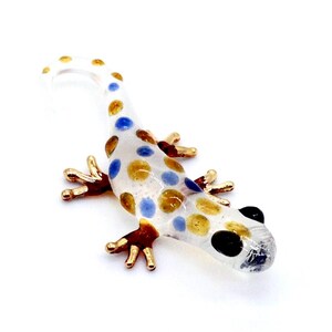 May include: A clear glass lizard figurine with gold, blue, and yellow polka dots. The lizard has gold-colored feet and a curled tail. The figurine is a decorative item.