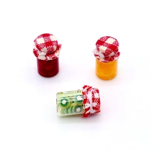 Könnte beinhalten: Drei Miniaturgläser mit Konfitüre. Zwei Gläser stehen aufrecht, eines gefüllt mit einer roten Substanz und das andere mit einer gelben Substanz. Das dritte Glas liegt horizontal und ist mit grünen Kiwischeiben gefüllt. Jedes Glas ist mit einem rot-weiß karierten Stoff bedeckt.