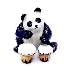 May include: A black and white ceramic panda figurine playing a pair of small, colorful drums. The panda is sitting on its haunches with its paws raised as if playing the drums.