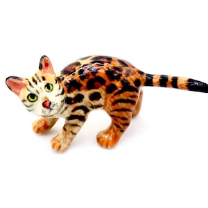 Könnte beinhalten: Eine Keramikfigur einer Bengal-Katze. Die Katze hat ein geflecktes Fell in Braun- und Schwarztönen, ein weißes Gesicht mit schwarzen Streifen, grüne Augen, rosa Ohren und eine rosa Nase. Die Katze befindet sich in einer spielerischen Pose.