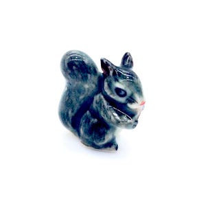 May include: A small, gray ceramic squirrel figurine with a pink nose. The squirrel is sitting on its haunches with its tail curled around its body.