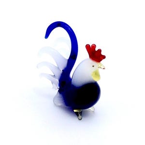 May include: A miniature glass rooster ornament. The rooster features a blue tail and a red comb. The body is white and blue, with a yellow beak. The ornament is set on a white surface.