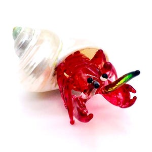 May include: A detailed glass figurine of a hermit crab. The crab is a vibrant red colour, with black eyes and a green and gold claw. It is housed in a pearlescent white and silver shell, set against a white background.