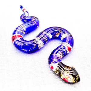 May include: A blue glass snake figurine with gold and red accents. The snake is curled in an S shape with its head facing the right.