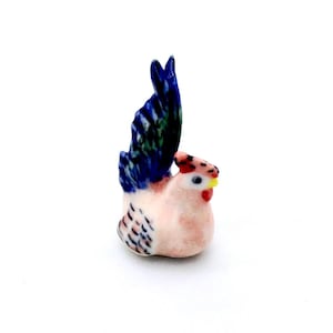 May include: A miniature ceramic rooster figurine. The rooster features a pink body, a red comb, and a yellow beak. Its tail feathers are blue and green. The figurine is set against a white backdrop.