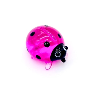 May include: A pink glass ladybug with black spots and black eyes. The ladybug is a small, decorative figurine.