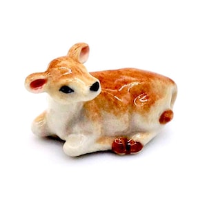 Op de afbeelding: Een keramieken beeldje van een kalf, liggend met het hoofd gedraaid. Het kalf is voornamelijk bruin en wit, met zwarte ogen en rode accenten op de oren en hoeven. De achtergrond is wit.