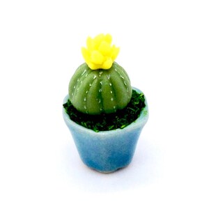 May include: A small green cactus with a yellow flower in a blue ceramic pot. The cactus is a miniature replica of a real cactus.