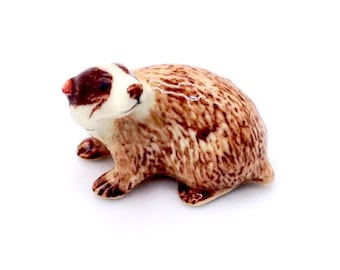 Może przedstawiać: Ceramiczna figurka borsuka z białą twarzą, brązowo-beżowym korpusem i ciemnobrązowymi oznaczeniami. Małe zwierzę jest ustawione na czterech łapach, z głową lekko obróconą na bok. Figurka jest na białym tle.