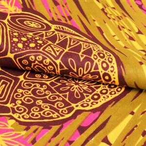 May include: Close-up of a folded fabric with a vibrant, abstract pattern. The design features large, dark brown shapes with intricate yellow outlines, set against a backdrop of olive green, yellow, and pink. The fabric has a textured appearance.