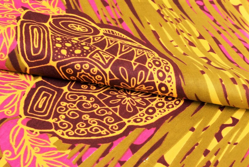 May include: Close-up of a folded fabric with a vibrant, abstract pattern. The design features large, dark brown shapes with intricate yellow outlines, set against a backdrop of olive green, yellow, and pink. The fabric has a textured appearance.