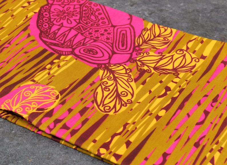 May include: Close-up of a folded fabric with a repeating pattern. The design features large, stylized floral and geometric shapes in shades of pink and brown, set against a mustard yellow background with diagonal stripes.