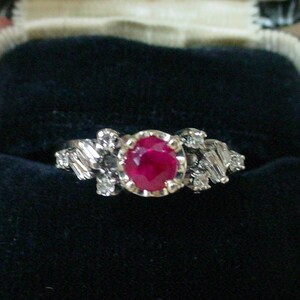 May include: A ring with a large, round, red ruby surrounded by smaller, clear diamonds. The ring is in a black velvet ring box.