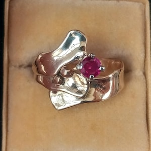 May include: A gold-toned ring with an abstract design and a round, red gemstone. The ring is set in a velvet-lined box, showcasing its intricate details and polished finish. The gemstone is a vibrant focal point.