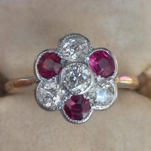 May include: A flower-shaped ring featuring a central diamond surrounded by six gemstones. The gemstones alternate between bright red rubies and clear diamonds, set in a gold band.