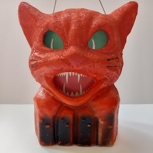 May include: A vintage Halloween cat decoration. The cat is red with green eyes and a wide, toothy grin. Black accents highlight the whiskers and the base of the cat. A black wire is attached for hanging. The cat is made of a textured material.