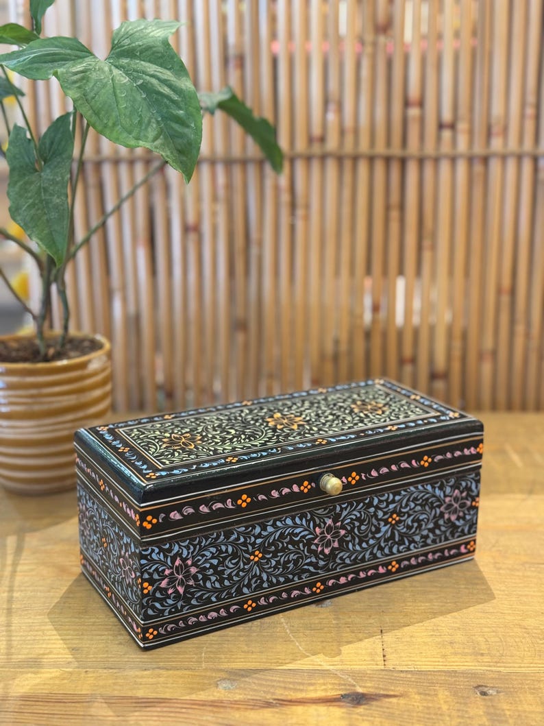 May include: A black wooden box with a floral design painted on the lid and sides. The box has a brass knob and is sitting on a wooden surface.
