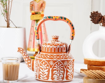 May include: A hand-painted orange kettle with white floral designs. The kettle has a pink handle with green accents and a white lid. The kettle is sitting on a white tablecloth.