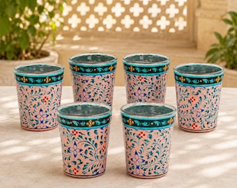 Puede incluir: Un juego de seis vasos con una base de color rosa claro, decorados con intrincados patrones de enredaderas en color verde azulado pintados a mano, pequeños detalles florales rojos y una banda horizontal oscura con motivos de flores naranjas.