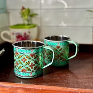 Puede incluir: Dos tazas decorativas de metal con fondo turquesa y estampados florales en rojo, amarillo y verde. Las tazas tienen interior plateado y asas turquesas a juego. Las tazas están sobre una bandeja de madera.