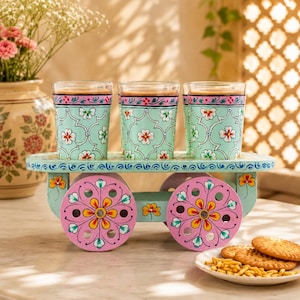 May include: Three cylindrical glass tumblers with a light blue floral pattern sit on a decorative wooden cart stand. The stand features two large pink wheels with yellow and white floral accents and a light blue base with matching floral motifs.