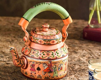 May include: A hand-painted pink metal teapot with a green handle and floral designs. The teapot features a scene of people riding horses and in a carriage. The teapot is sitting on a wooden surface with two blue and white teacups.