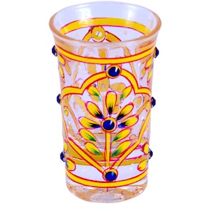 May include: A clear glass tumbler featuring hand-painted yellow and pink floral patterns with blue accents. The cylindrical vessel is decorated with small blue glass beads attached to the exterior sides, creating a textured and colorful appearance.