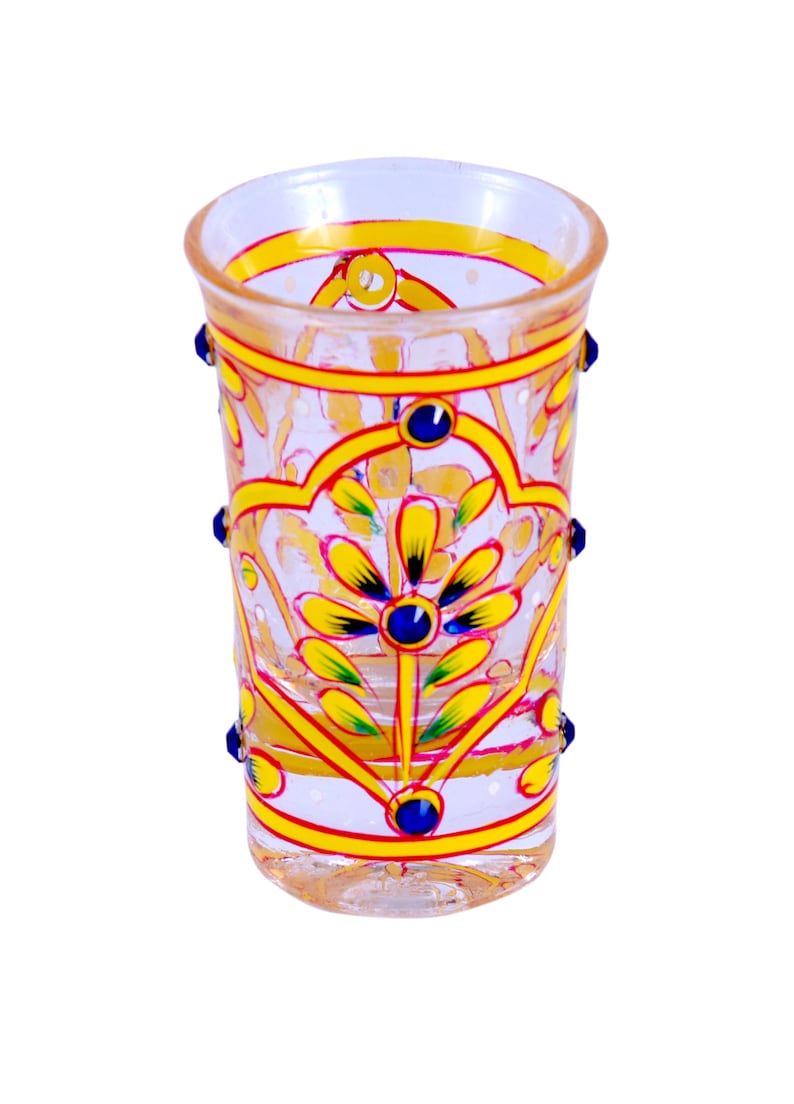 May include: A clear glass tumbler featuring hand-painted yellow and pink floral patterns with blue accents. The cylindrical vessel is decorated with small blue glass beads attached to the exterior sides, creating a textured and colorful appearance.