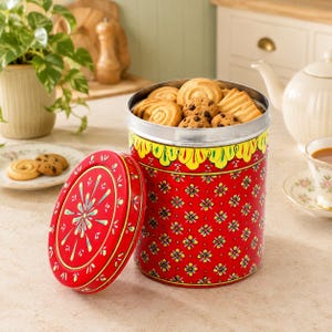 May include: A cylindrical red metal storage tin featuring a repeating yellow and green floral pattern and a decorative yellow scalloped border at the top. The lid is detached and displays a central starburst design. The tin is filled with assorted cookies.